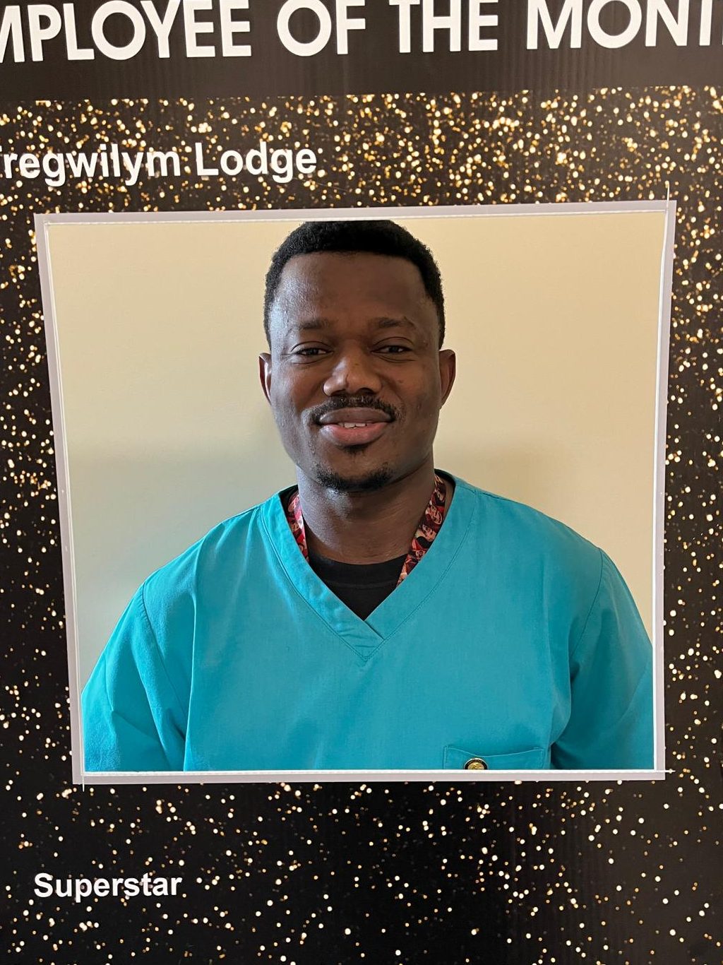 Portrait of a man wearing a turquoise scrub top, smiling, in front of a golden sparkly background with the text 'EMPLOYEE OF THE MONTH' and his name, Gregwilym Lodge.