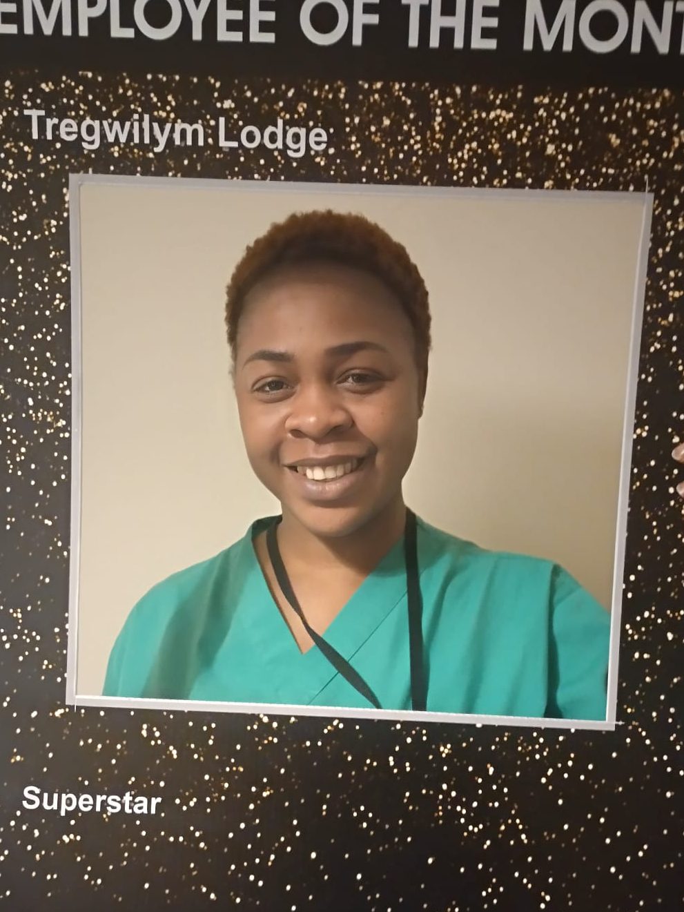 A smiling woman wearing green scrubs stands in front of a black and gold 'Employee of the Month' plaque, which features her name, Tregwilym Lodge, and the word 'Superstar'.